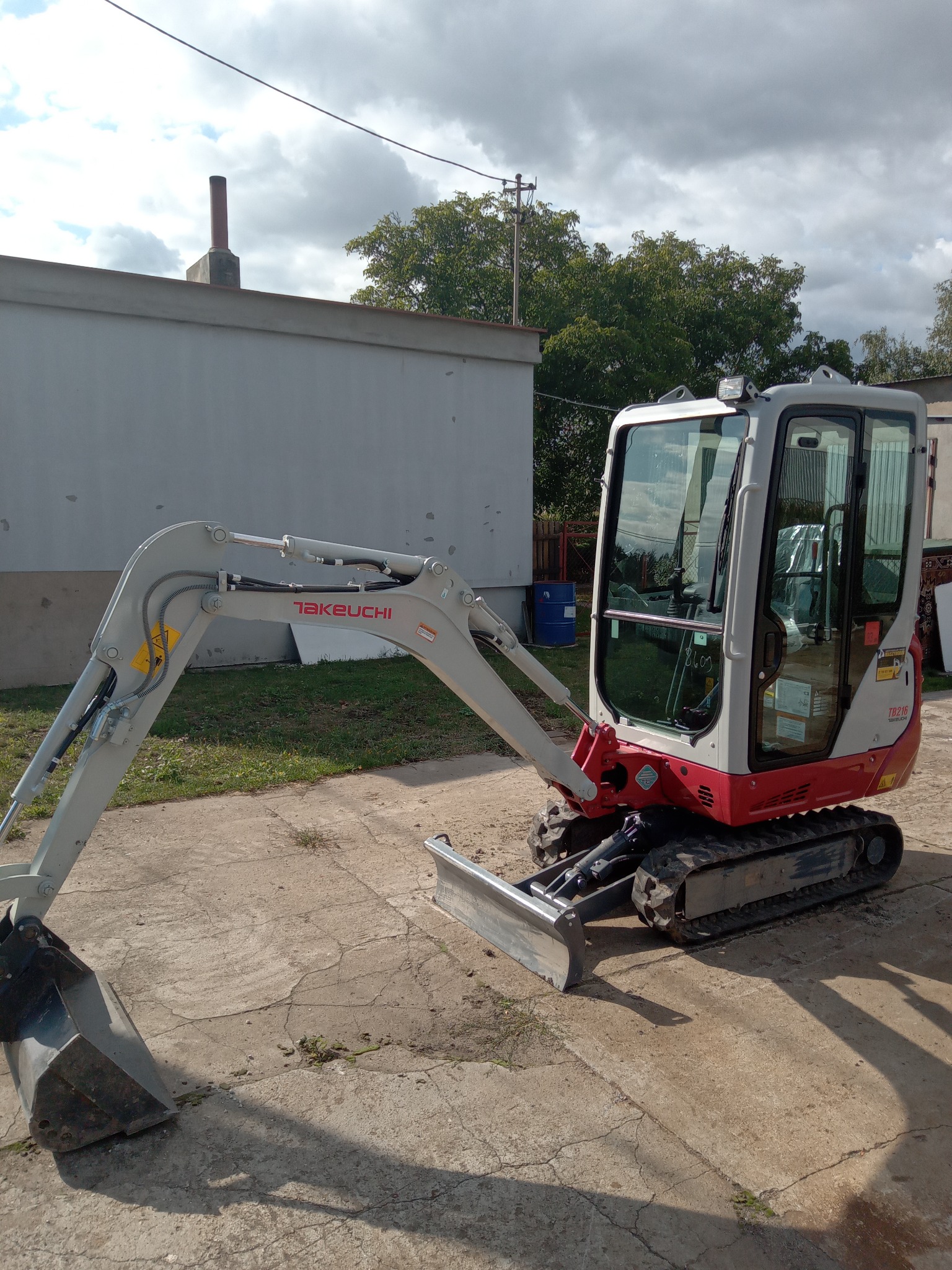 Kompaktowa minikoparka Takeuchi TB216 z łyżką i lemieszem, zaparkowana na betonowym podłożu, widok z boku, w tle budynek i drzewa.