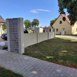 Nowoczesne ogrodzenie z betonowych bloczków i gładkiego betonu, oddzielające posesję z trawnikiem i kostką brukową, w tle widoczny dom z żółtą elewacją pod błękitnym niebem.