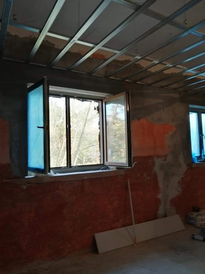 Wnętrze w trakcie remontu: okna częściowo otwarte, ściany w surowym tynku, widoczna metalowa konstrukcja sufitu podwieszanego.