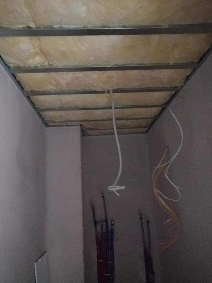 Sufit podwieszany z izolacją z wełny mineralnej w trakcie montażu, widoczne kable elektryczne i rury instalacyjne na ścianie.