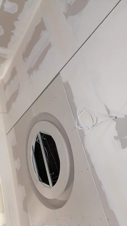 Ściana z zamontowanym okrągłym oknem, w trakcie prac wykończeniowych, widoczne ślady po szpachlowaniu i wystający kabel elektryczny.
