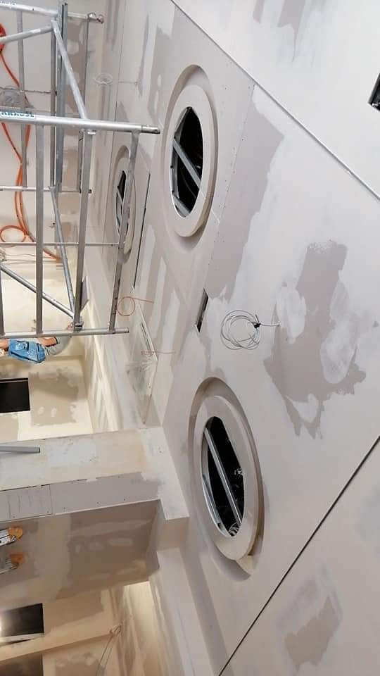 Ściana w trakcie prac wykończeniowych z okrągłymi otworami, widoczne ślady po szpachlowaniu, kabel wystający ze ściany i rusztowanie.