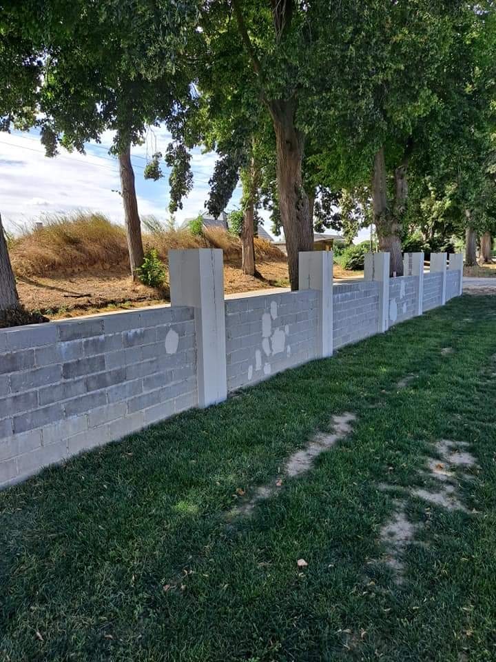 Murek z szarej cegły betonowej z białymi słupkami, widoczne ślady po szpachlowaniu, otoczony zielonym trawnikiem i drzewami.