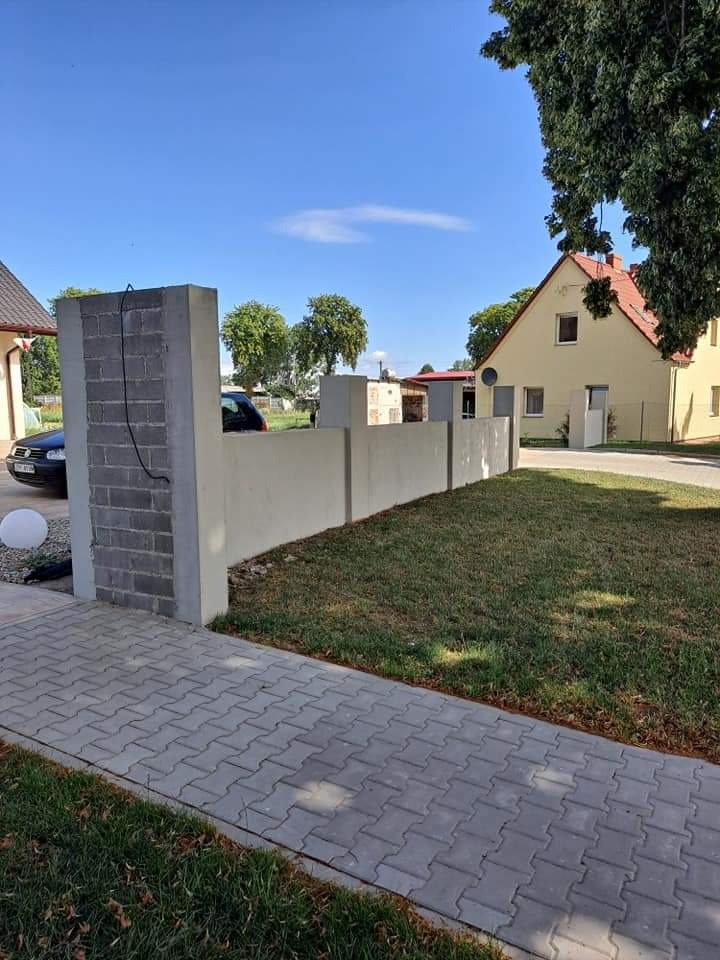 Nowoczesne ogrodzenie z betonowych bloczków i gładkiego betonu, oddzielające posesję z trawnikiem i kostką brukową, w tle widoczny dom z żółtą elewacją pod błękitnym niebem.