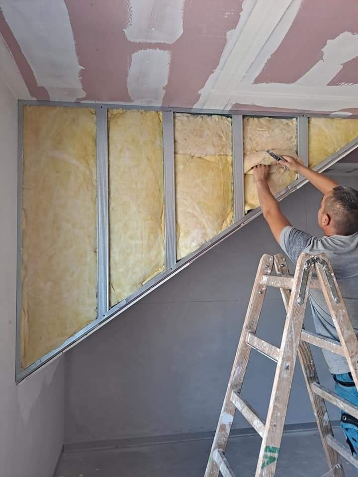 Montaż izolacji z wełny mineralnej w metalowym stelażu pod skosem poddasza, widoczna drabina i częściowo wyszpachlowany sufit.