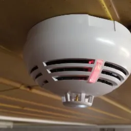 Zamontowany na suficie czujnik dymu z czerwoną diodą sygnalizacyjną, widoczne elementy instalacji.