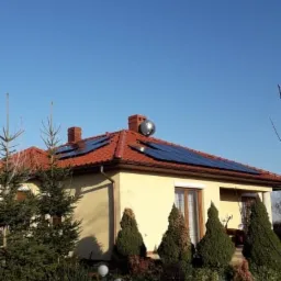 Dom jednorodzinny z panelami słonecznymi na czerwonym dachu, widoczny z poziomu ogrodu w słoneczny dzień. Na dachu zainstalowany jest również kolektor słoneczny.