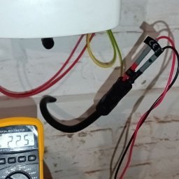 Tania firma z Ukrainy posiadająca Niemieckie doświadczenie - Pomiar napięcia za pomocą multimetru na instalacji elektrycznej z czerwonymi i czarnymi przewodami na tle ceglanej ściany.