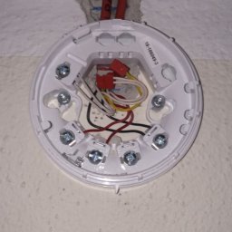 Tania firma z Ukrainy posiadająca Niemieckie doświadczenie - Biała podstawa czujnika dymu z widocznymi przewodami i śrubami montażowymi, zamocowana do ściany wewnątrz budynku. Widoczne czerwone kable w otworze w ścianie.