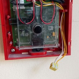 Tania firma z Ukrainy posiadająca Niemieckie doświadczenie - Czerwona obudowa centrali alarmowej z widocznymi przewodami, zielonymi złączkami i elementami elektroniki, zamontowana na jasnej ścianie.