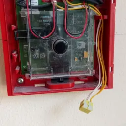 Czerwona obudowa centrali alarmowej z widocznymi przewodami, zielonymi złączkami i elementami elektroniki, zamontowana na jasnej ścianie.