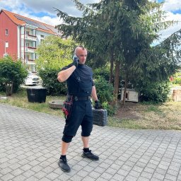 Tania firma z Ukrainy posiadająca Niemieckie doświadczenie - Elektryk w kombinezonie rozmawia przez telefon, trzymając skrzynkę narzędziową na tle budynku z panelami słonecznymi na dachu.