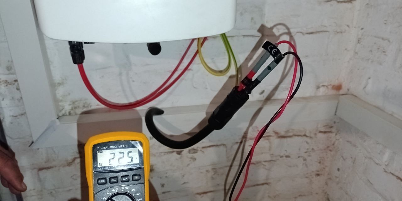 Pomiar napięcia za pomocą multimetru na instalacji elektrycznej z czerwonymi i czarnymi przewodami na tle ceglanej ściany.