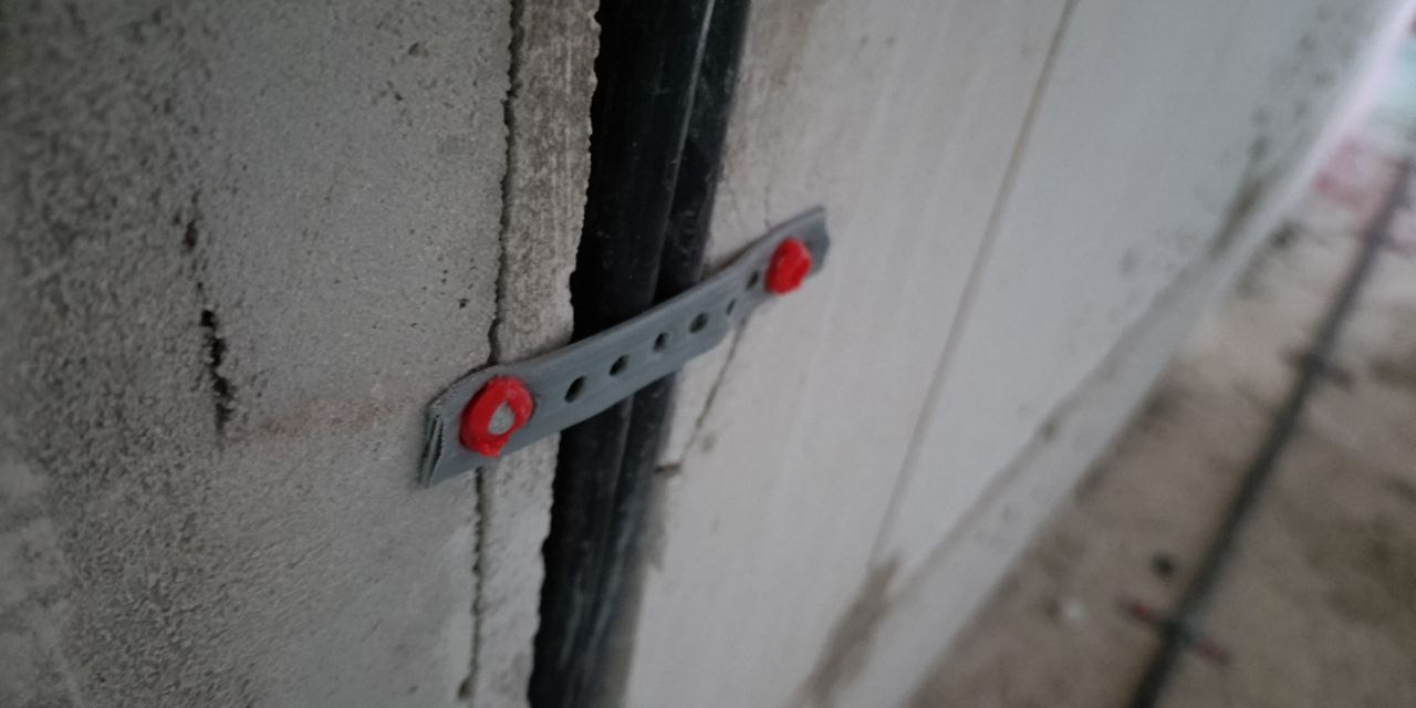 Przymocowane kable elektryczne do ściany z betonu komórkowego za pomocą szarej listwy z czerwonymi kołkami. Widoczne otwory w listwie i faktura ściany.