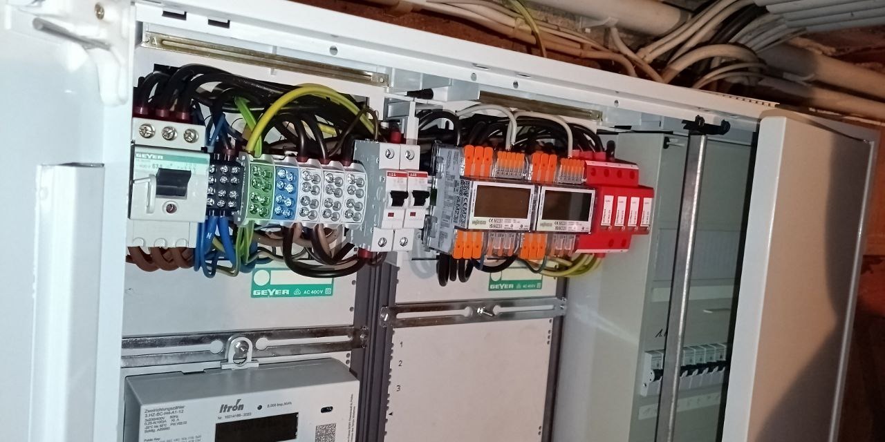 Otwarta skrzynka elektryczna z zabezpieczeniami, przewodami i licznikiem Itron. Widoczne elementy marki Geyer. Układ kabli i komponentów wewnątrz szafy.