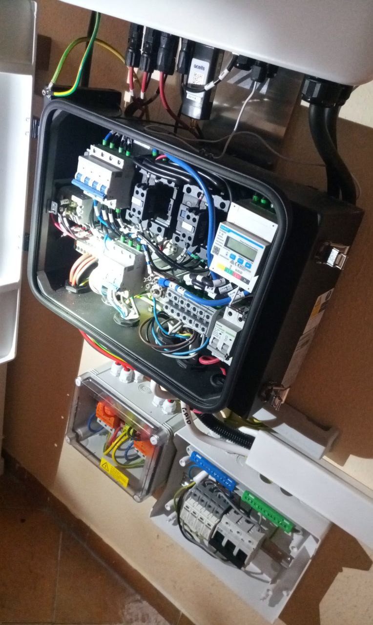 Otwarta skrzynka z okablowaniem i zabezpieczeniami instalacji fotowoltaicznej, widoczne przewody, bezpieczniki i licznik. Dolne puszki z elementami elektrycznymi.