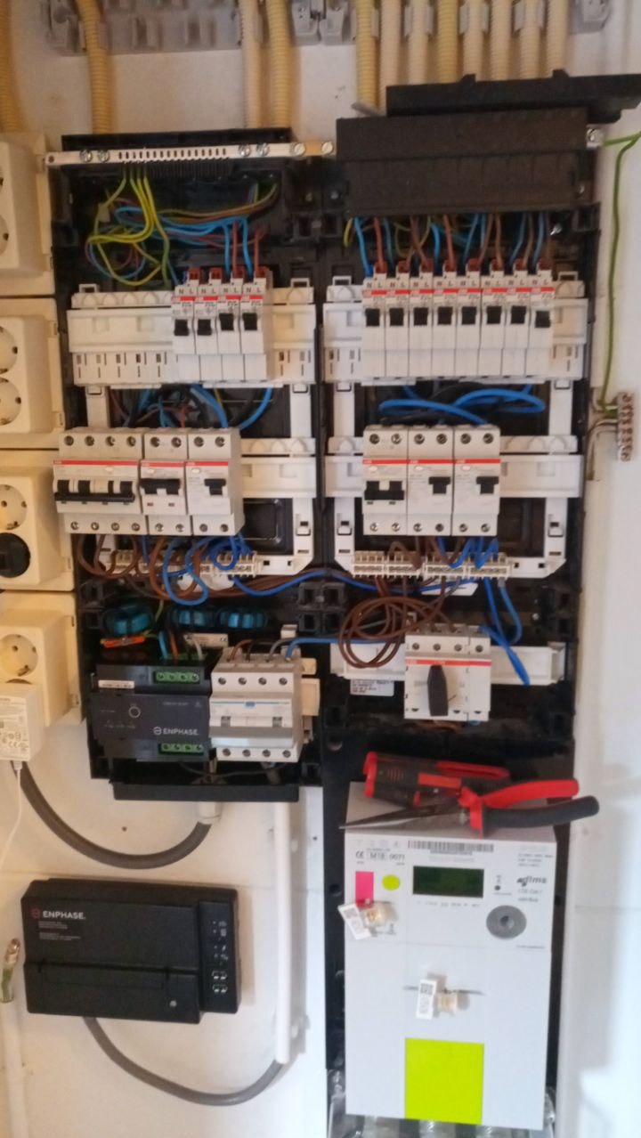 Rozdzielnica elektryczna z bezpiecznikami, przewodami i licznikiem energii, uzupełniona o narzędzia. Widoczne elementy systemu Enphase.