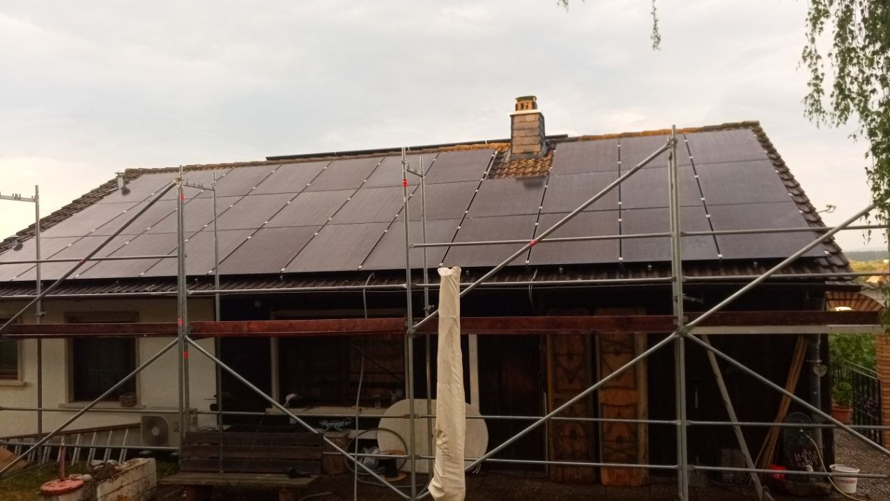 Dom z panelami fotowoltaicznymi na dachu, częściowo zasłonięty rusztowaniem. Widoczny komin i fragmenty elewacji. Pochmurne niebo w tle.
