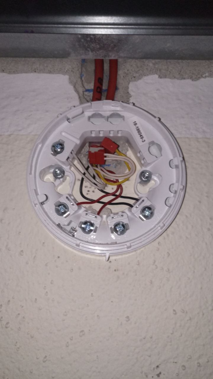 Biała podstawa czujnika dymu z widocznymi przewodami i śrubami montażowymi, zamocowana do ściany wewnątrz budynku. Widoczne czerwone kable w otworze w ścianie.