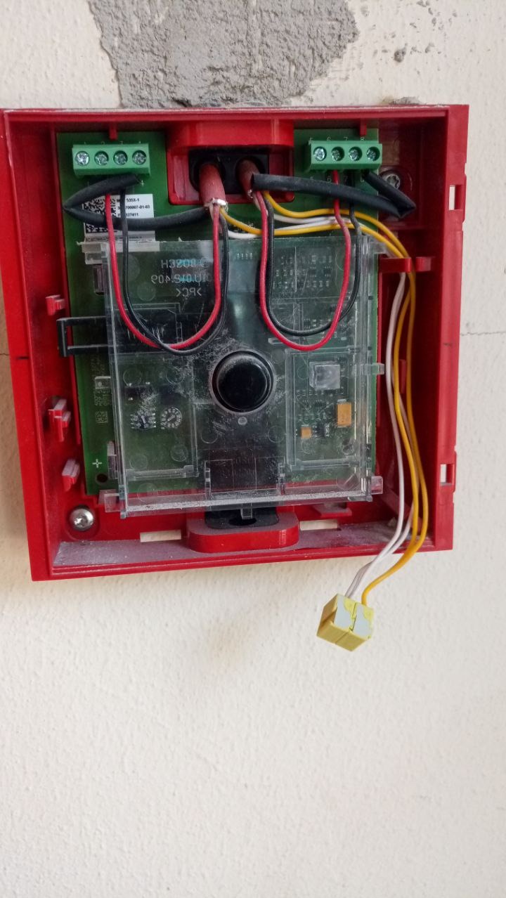 Czerwona obudowa centrali alarmowej z widocznymi przewodami, zielonymi złączkami i elementami elektroniki, zamontowana na jasnej ścianie.