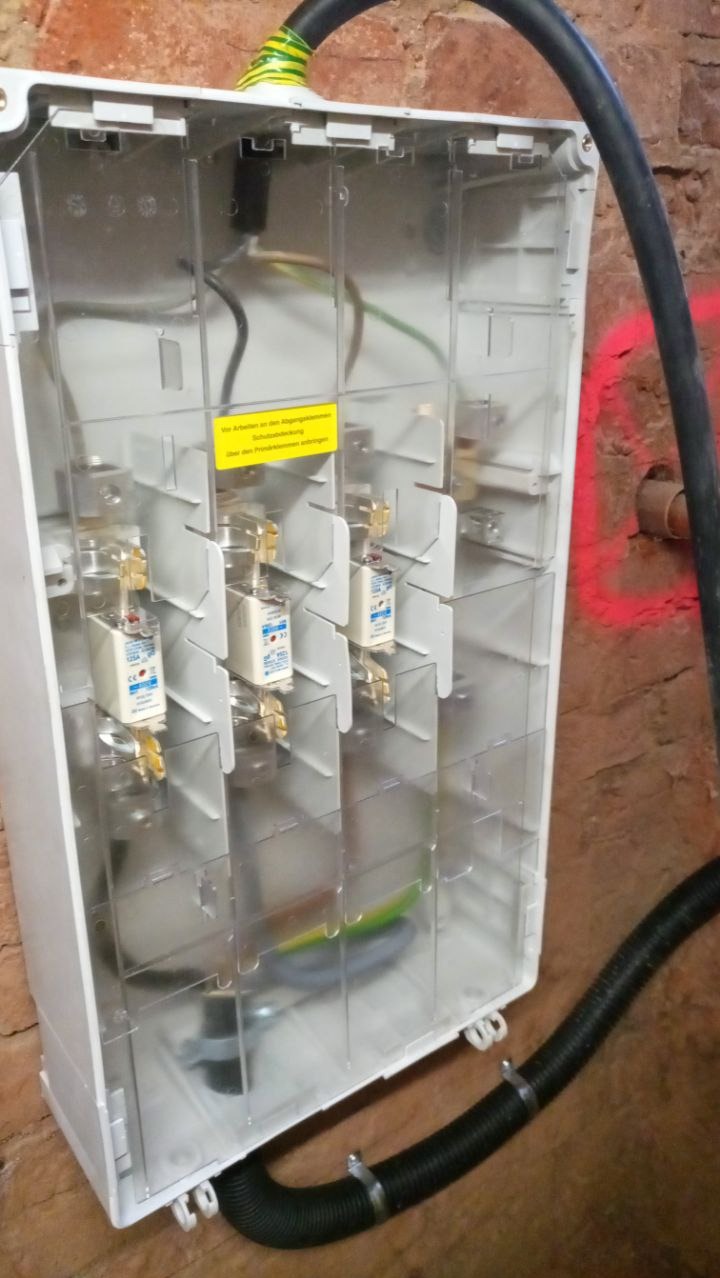 Rozdzielnica elektryczna z bezpiecznikami topikowymi w przezroczystej obudowie, widoczne przewody, zamontowana na ceglanej ścianie. Czarna rura karbowana u dołu.