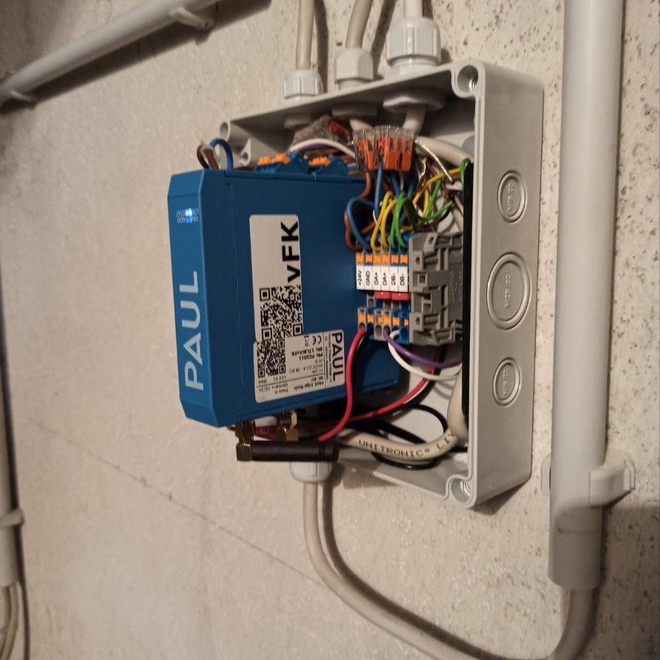 Rozdzielnica elektryczna z modułem PAUL VFK, widoczne okablowanie i złącza. Szara puszka instalacyjna zamontowana na ścianie, przewody w białych peszlach.