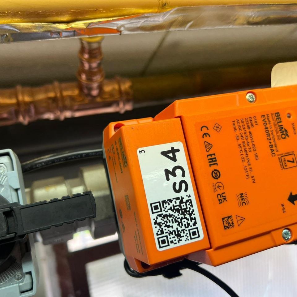 Pomarańczowy siłownik Belimo EVO40R2+BAC z kodem QR i oznaczeniem S34, zamontowany na instalacji. Widoczne miedziane rury i informacje techniczne na obudowie.