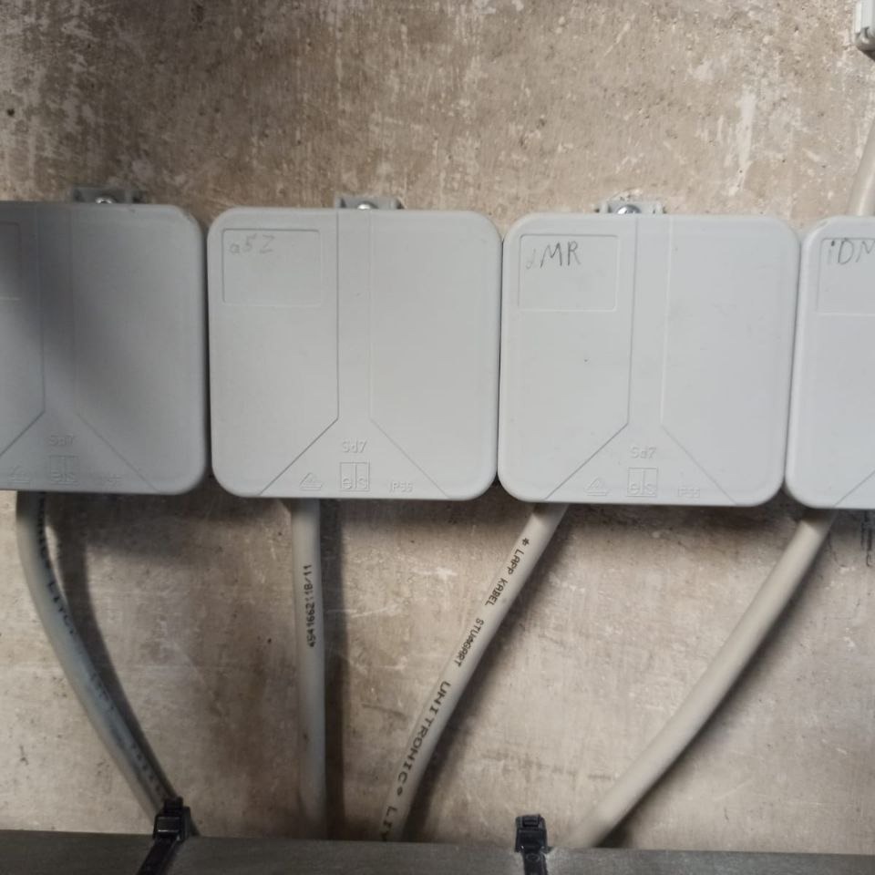 Szereg szarych puszek instalacyjnych z kablami na szorstkiej, betonowej ścianie. Na puszkach widoczne odręczne napisy: a52, JMR, DM.