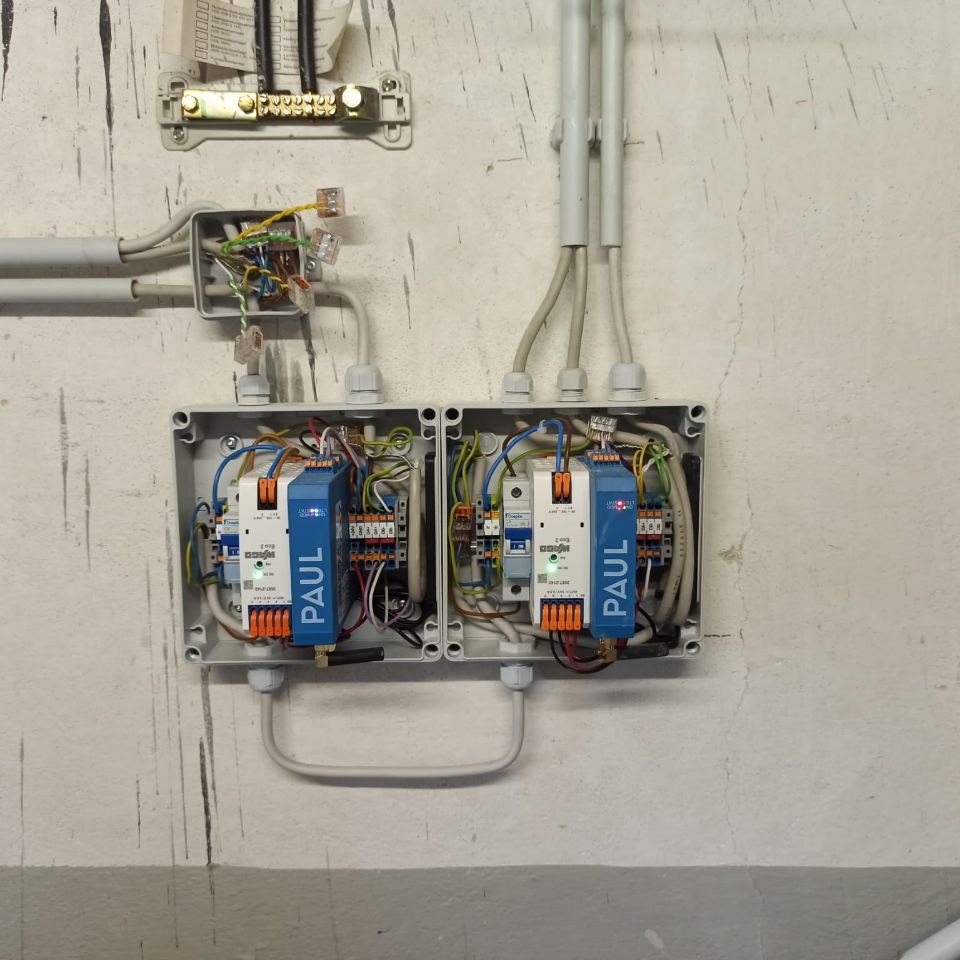 Dwa szare panele sterowania z niebieskimi modułami PAUL, zamontowane na ścianie z widocznymi przewodami. Górna część ściany z dodatkowymi elementami instalacji.