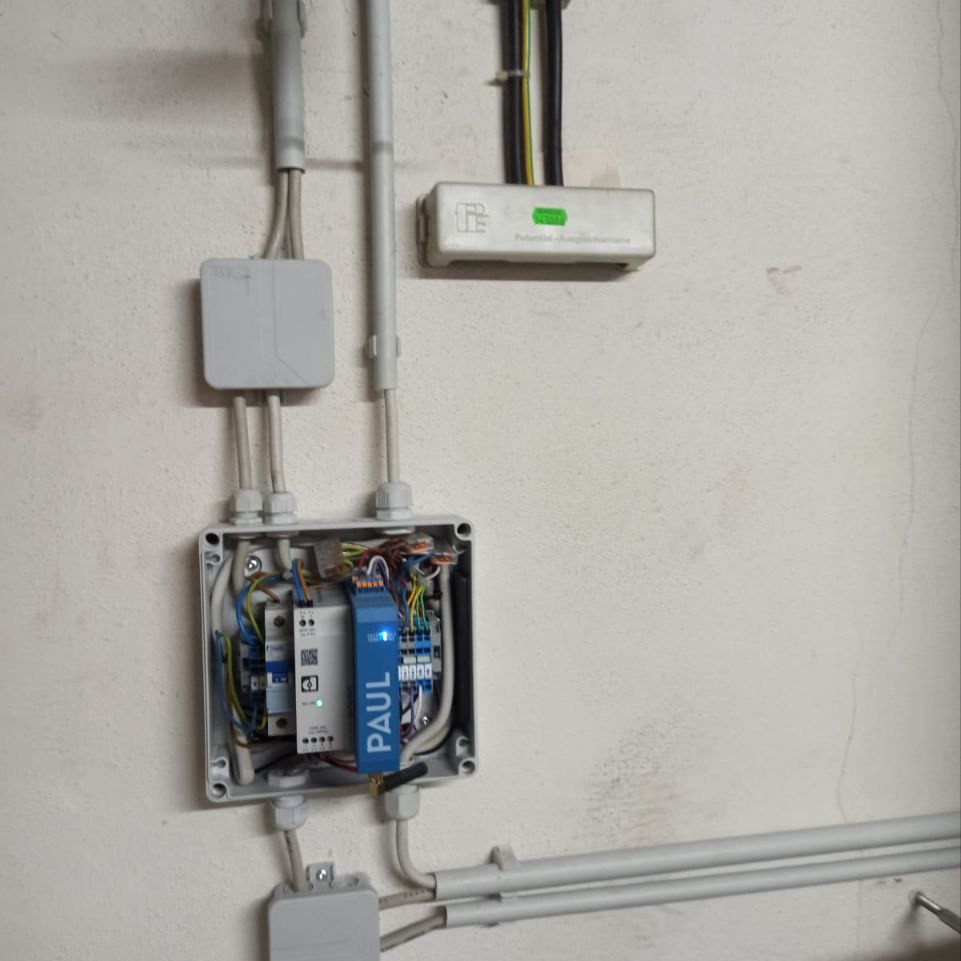 Otwarta puszka instalacyjna z widocznymi przewodami, modułami i listwami zaciskowymi marki Paul, zamontowana na ścianie. Widoczne peszle i inne elementy instalacji.