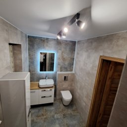 TupTech - Wykończona łazienka z szarymi płytkami, podświetlanym lustrem, białą umywalką i toaletą, oraz drewnianą ościeżnicą drzwiową.