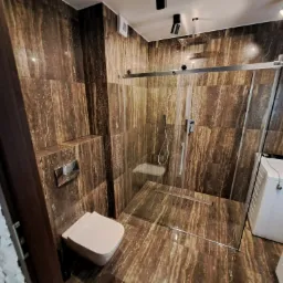Nowoczesna łazienka z kabiną prysznicową typu walk-in, wykończona brązowymi płytkami imitującymi kamień, z wiszącą toaletą i pralką w rogu.