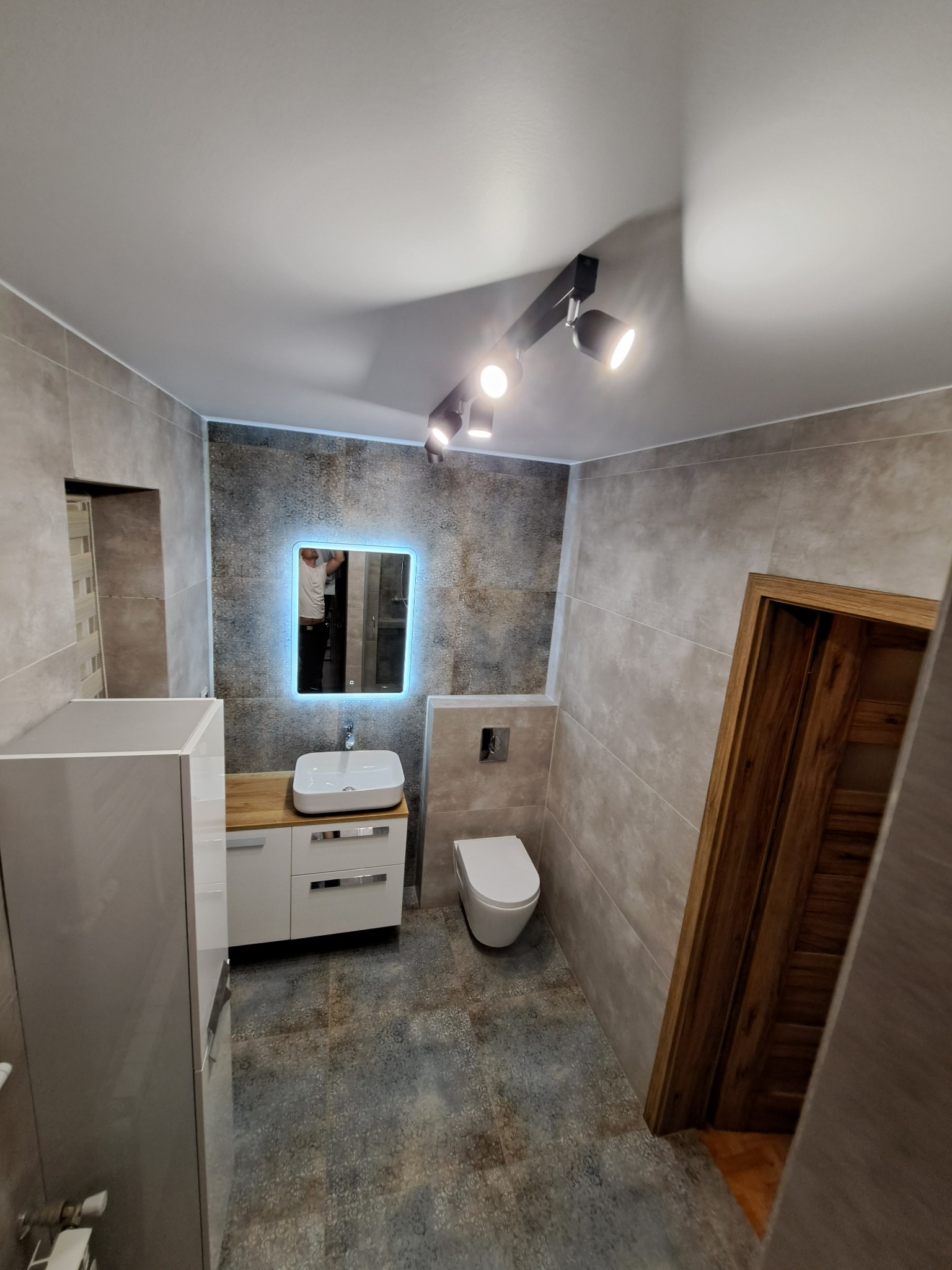 Wykończona łazienka z szarymi płytkami, podświetlanym lustrem, białą umywalką i toaletą, oraz drewnianą ościeżnicą drzwiową.