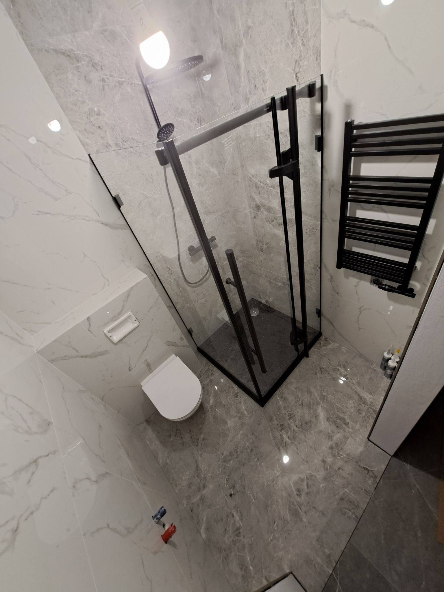 Nowoczesna łazienka z kabiną prysznicową typu walk-in z czarnymi profilami, grzejnikiem drabinkowym w kolorze czarnym, marmurowe płytki na ścianach i podłodze, wisząca toaleta.