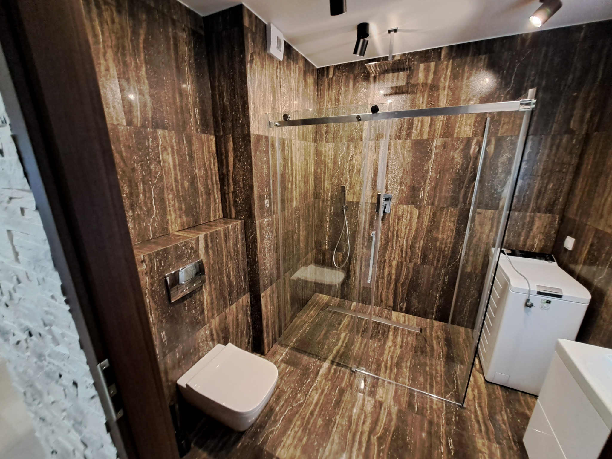Nowoczesna łazienka z kabiną prysznicową typu walk-in, wykończona brązowymi płytkami imitującymi kamień, z wiszącą toaletą i pralką w rogu.