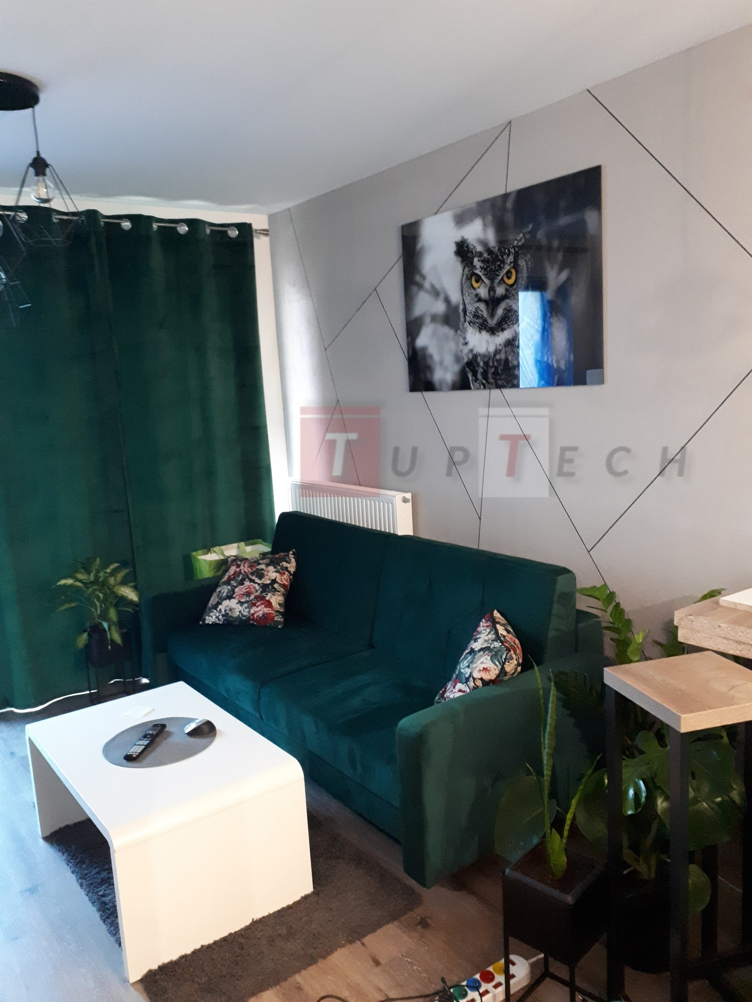 Salon z zieloną, welurową sofą, białym stolikiem kawowym i dekoracyjną ścianą z motywem geometrycznym oraz obrazem sowy.