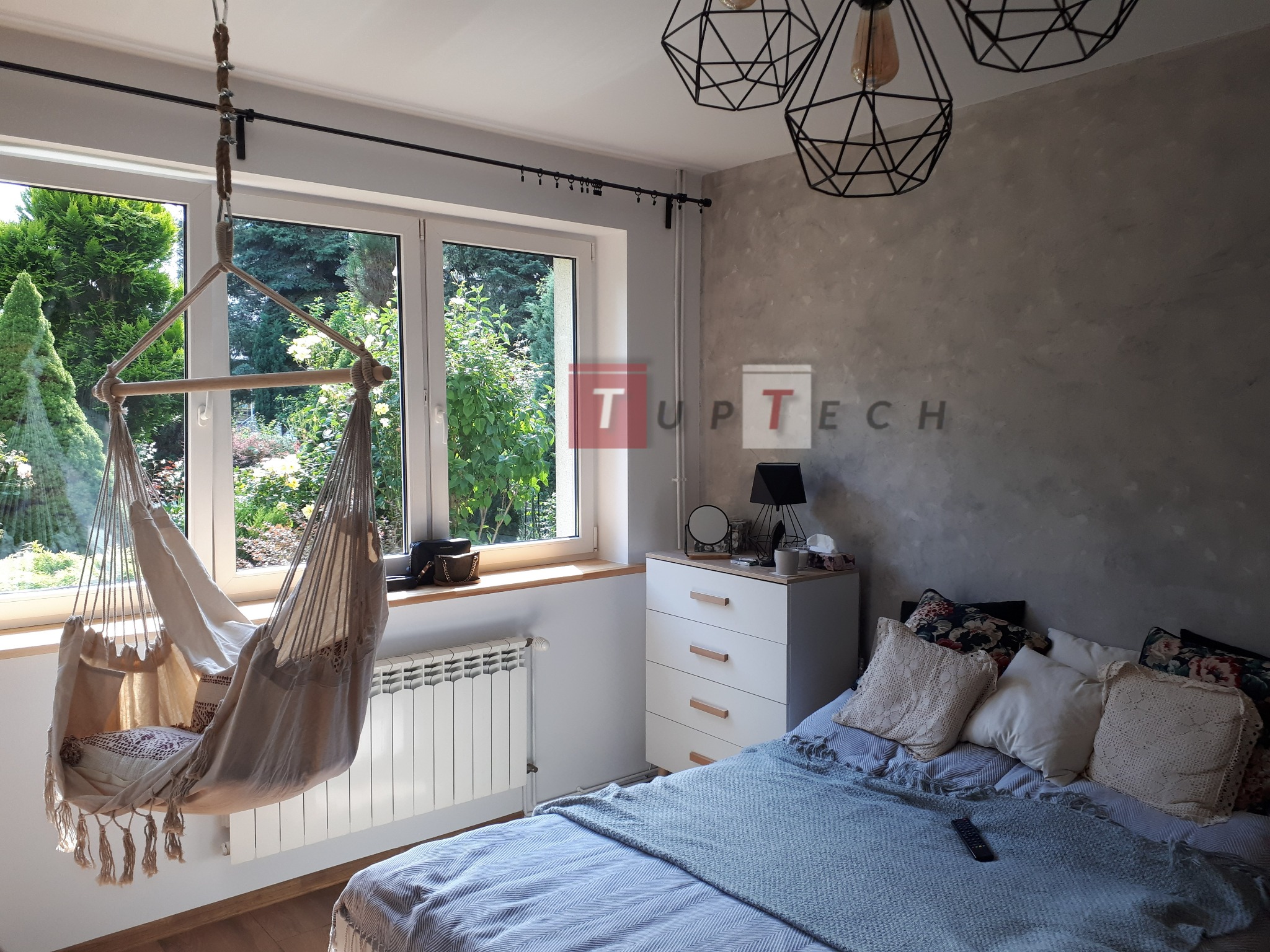 Jasny pokój z wiszącym fotelem hamakowym przy oknie, widok na zieleń za oknem, dekoracyjna lampa sufitowa i szara ściana w tle.