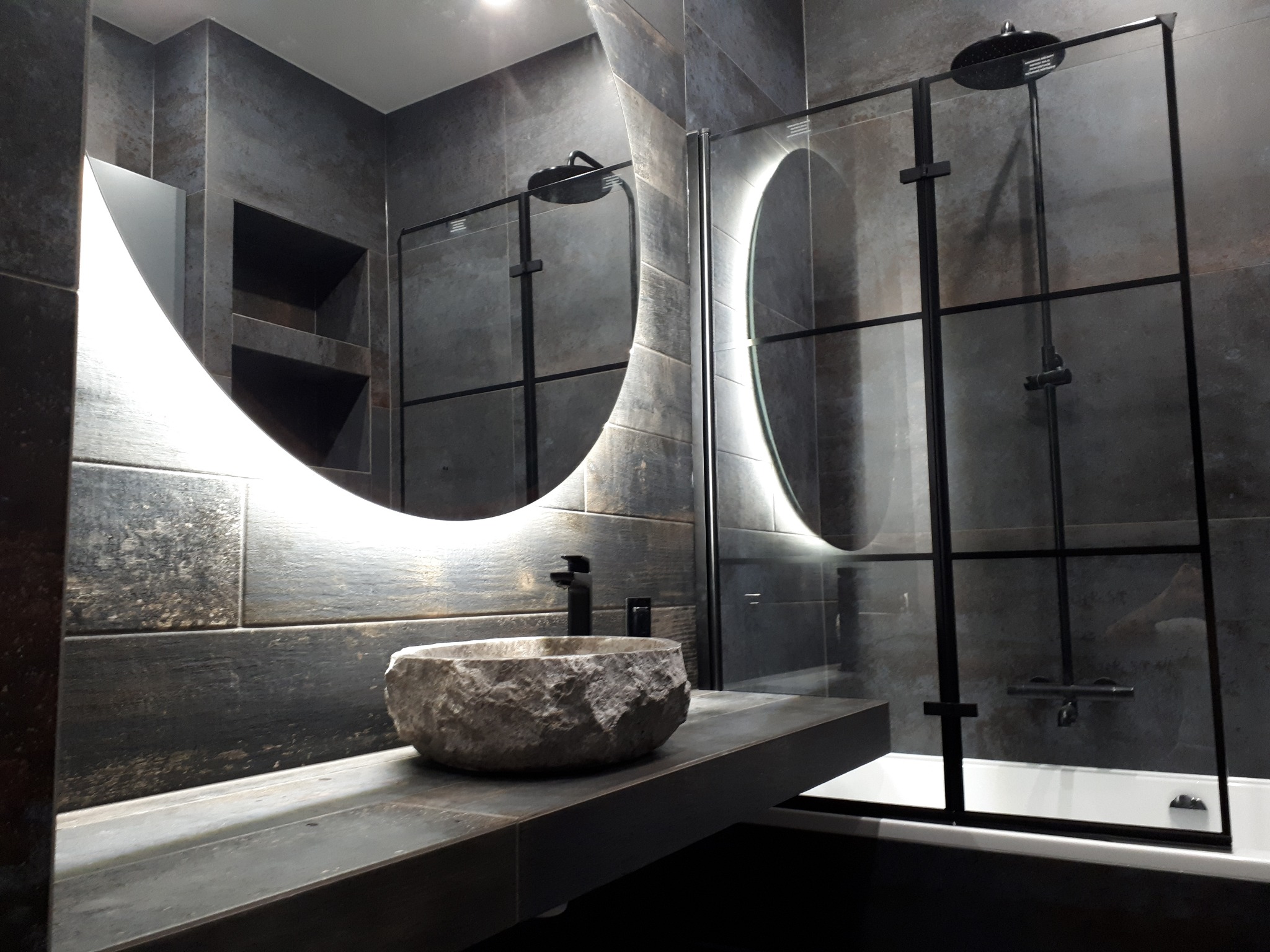 Nowoczesna łazienka z szarą, teksturowaną okładziną ścienną, okrągłym lustrem z podświetleniem LED, kamienną umywalką i kabiną prysznicową z czarnymi profilami.