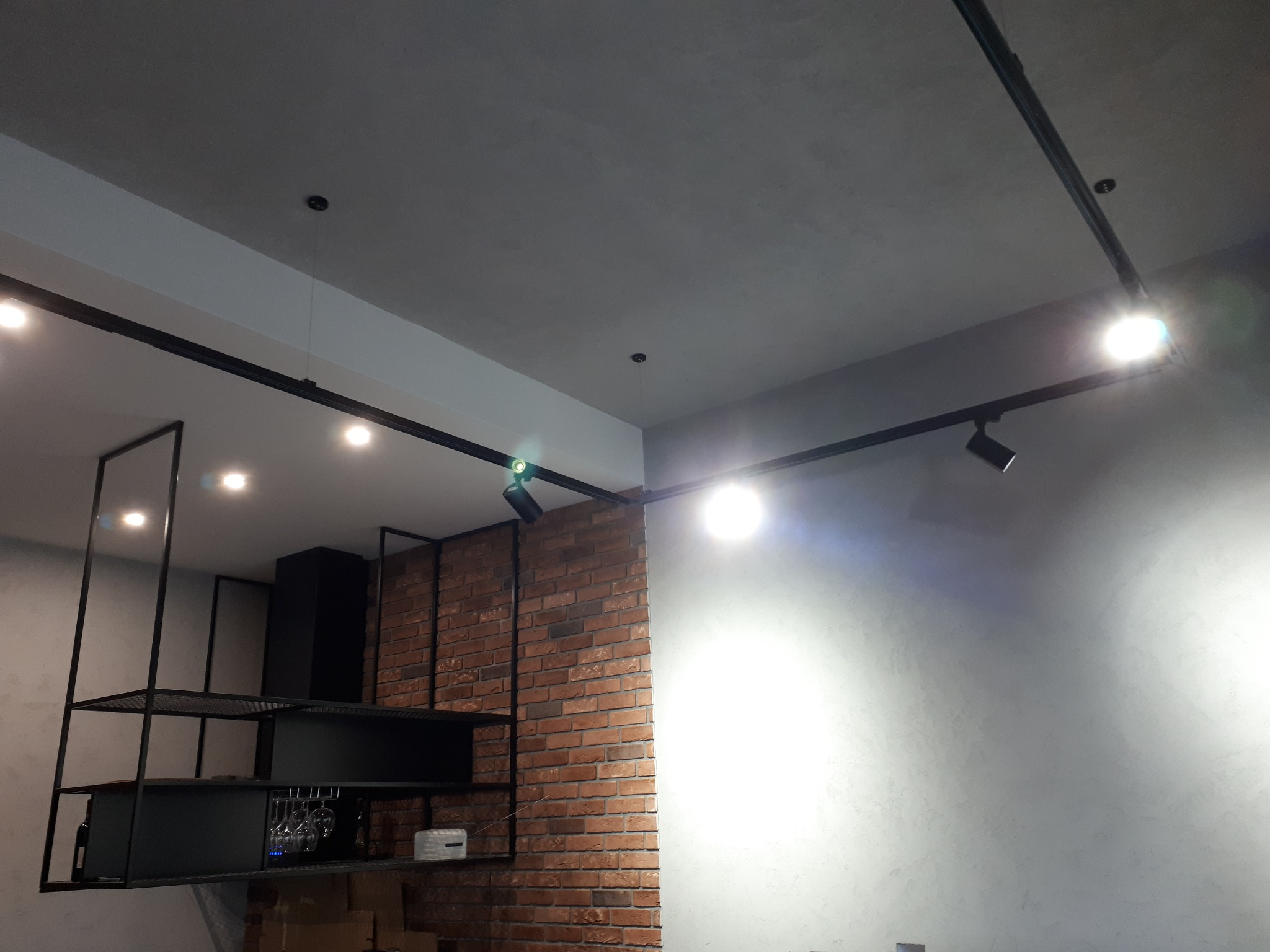 Wnętrze z oświetleniem szynowym, fragmentem ceglanej ściany, metalowym regałem z czarnymi elementami i szklankami na wino.