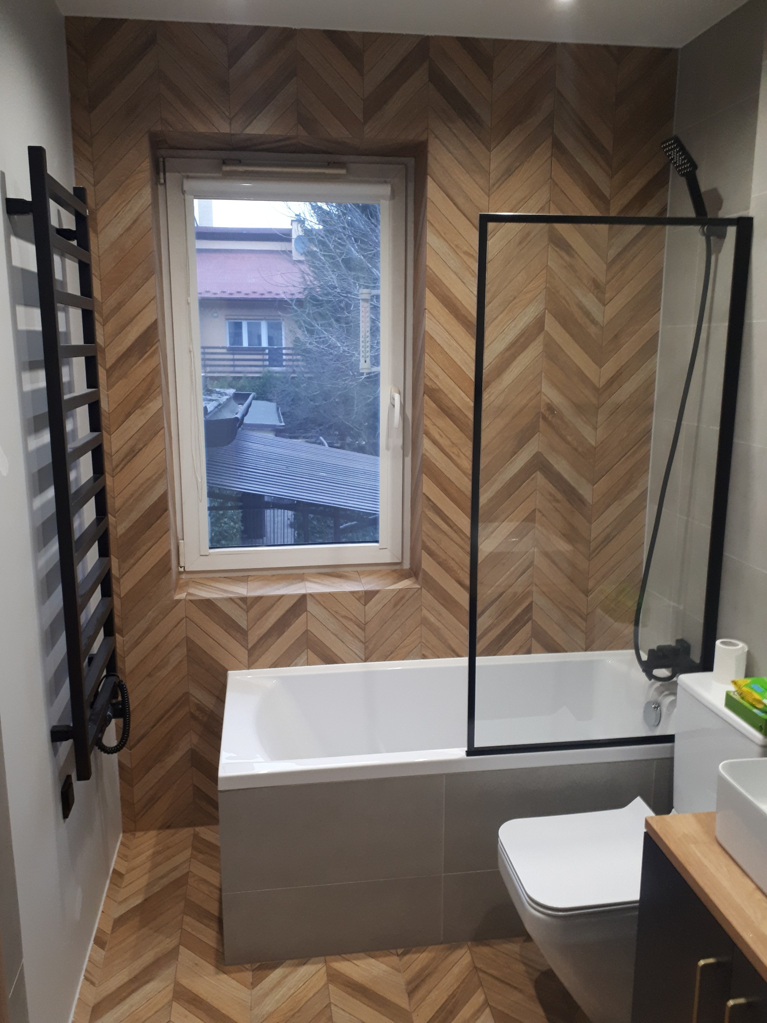 Nowoczesna łazienka z drewnopodobną okładziną ułożoną w jodełkę na ścianach i podłodze, kabina prysznicowa z czarną ramą, grzejnik drabinkowy i widok z okna na dom.