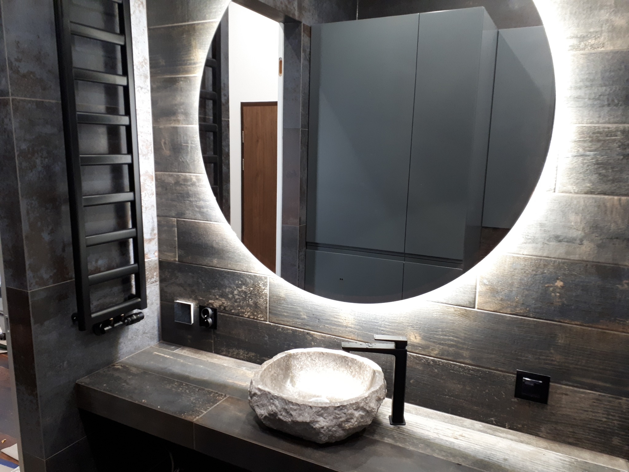Wykończona łazienka z umywalką z kamienia, czarną armaturą, podświetlanym lustrem i grzejnikiem drabinkowym na ścianie z płytek imitujących drewno.