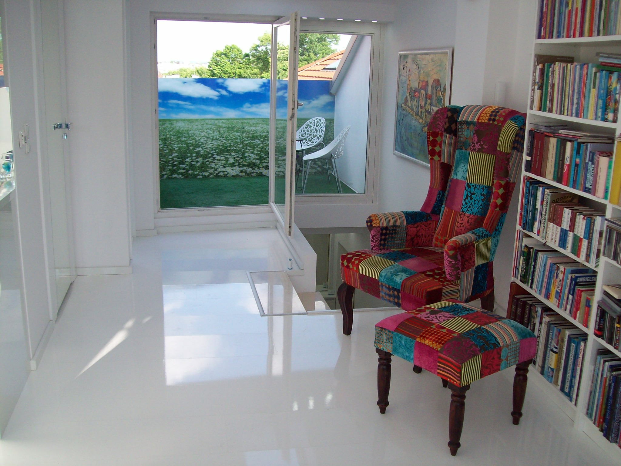 Jasne wnętrze z białymi ścianami i podłogą z żywicy, fototapeta z motywem łąki za oknem balkonowym, patchworkowy fotel uszak z podnóżkiem i regał z książkami.