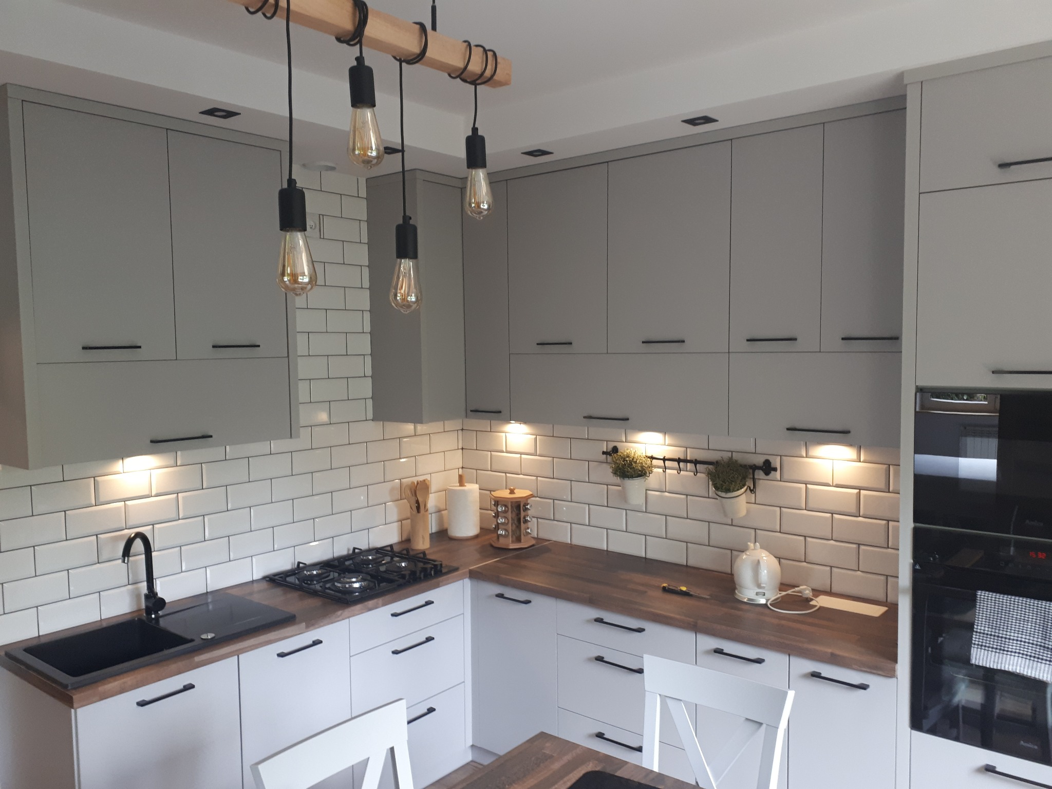 Jasna kuchnia z szarymi szafkami, białymi płytkami na ścianie, drewnianym blatem i trzema żarówkami wiszącymi na drewnianej belce.