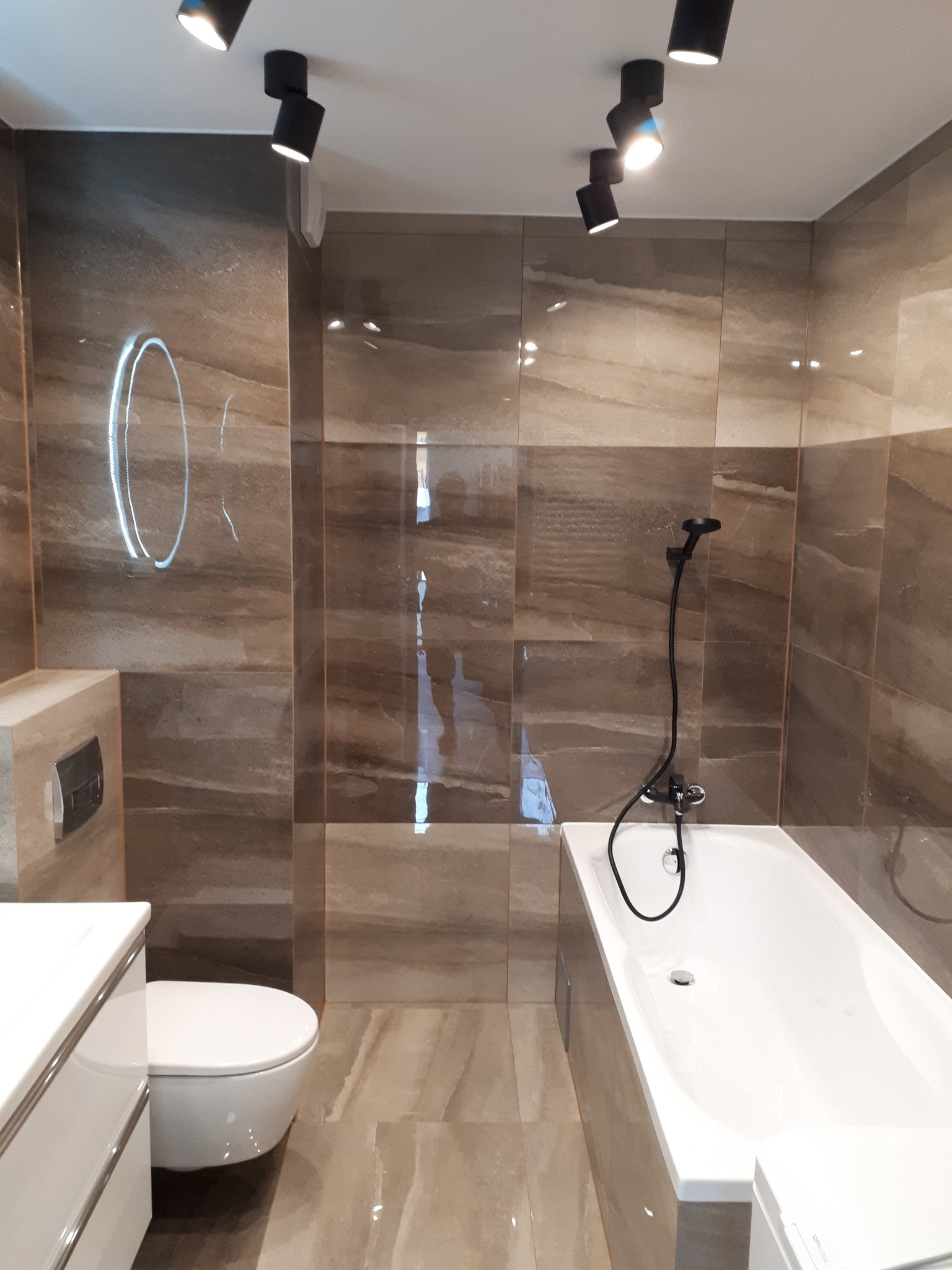 Nowoczesna łazienka z brązowymi płytkami imitującymi marmur, białą armaturą i czarną baterią prysznicową oraz okrągłym lustrem z podświetleniem LED.