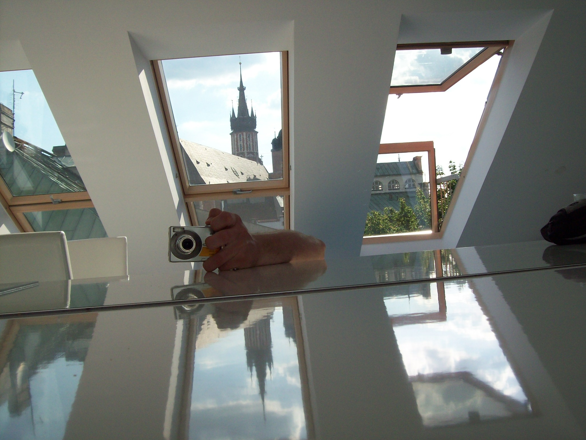 Poddasze z oknami dachowymi, widok na wieżę kościoła odbijający się w błyszczącej powierzchni, ręka trzymająca aparat fotograficzny.