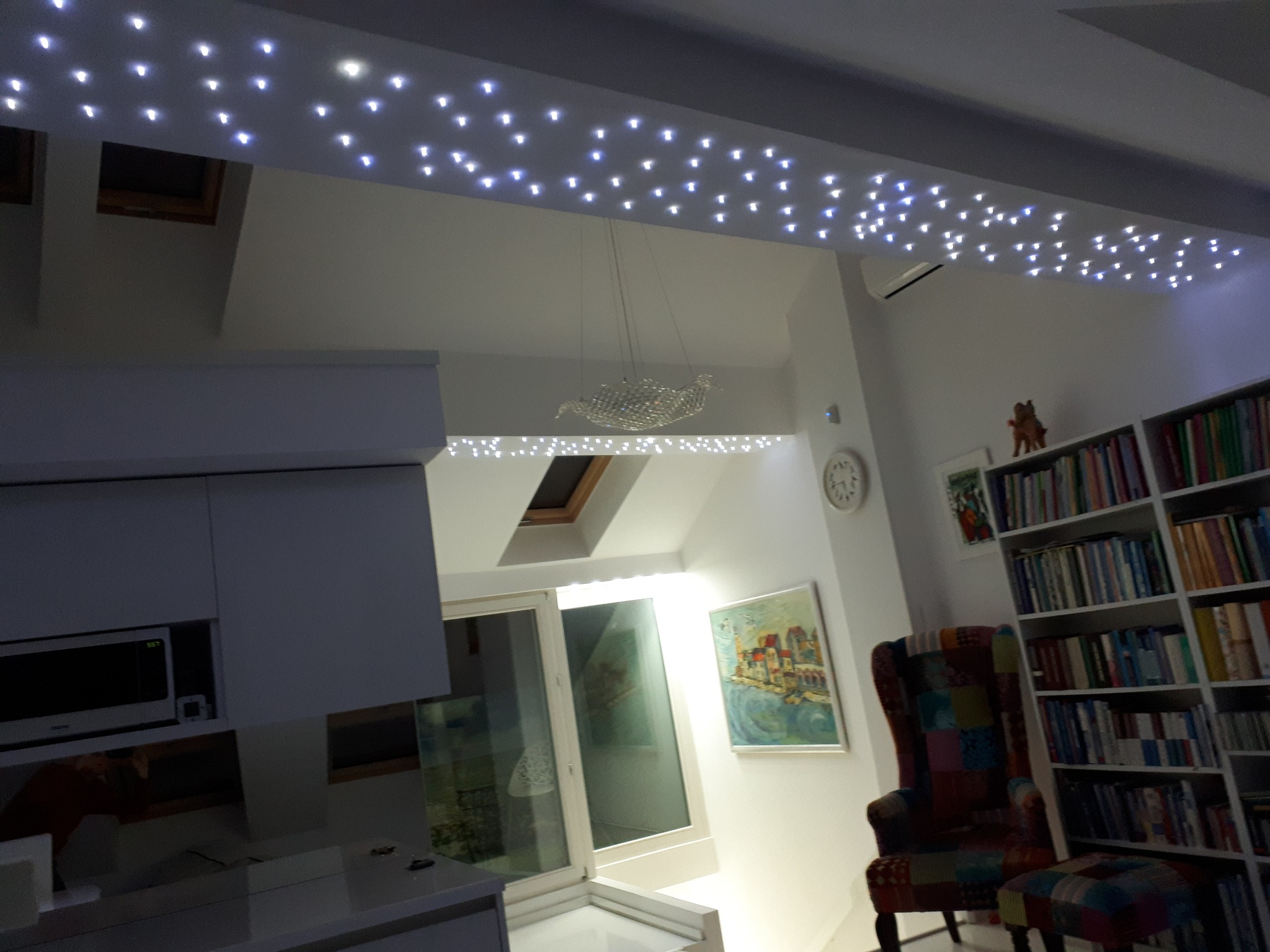 Salon z oświetleniem sufitowym imitującym gwiazdy, widoczna zabudowa okna dachowego, krzesło patchworkowe i regał z książkami.