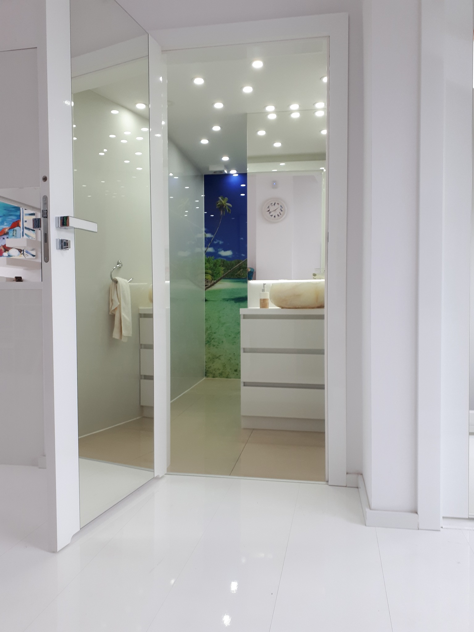 Nowoczesna, biała łazienka z szklanymi drzwiami i fototapetą z motywem tropikalnej plaży, widoczna umywalka nablatowa i szafka z szufladami.