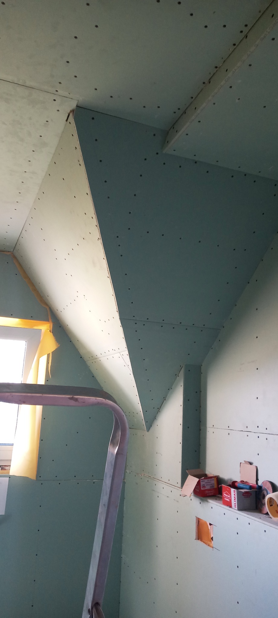 Skośne poddasze w trakcie obkładania płytami gipsowo-kartonowymi, widoczna drabina aluminiowa, otwór na instalację i pudełka z wkrętami na półce.
