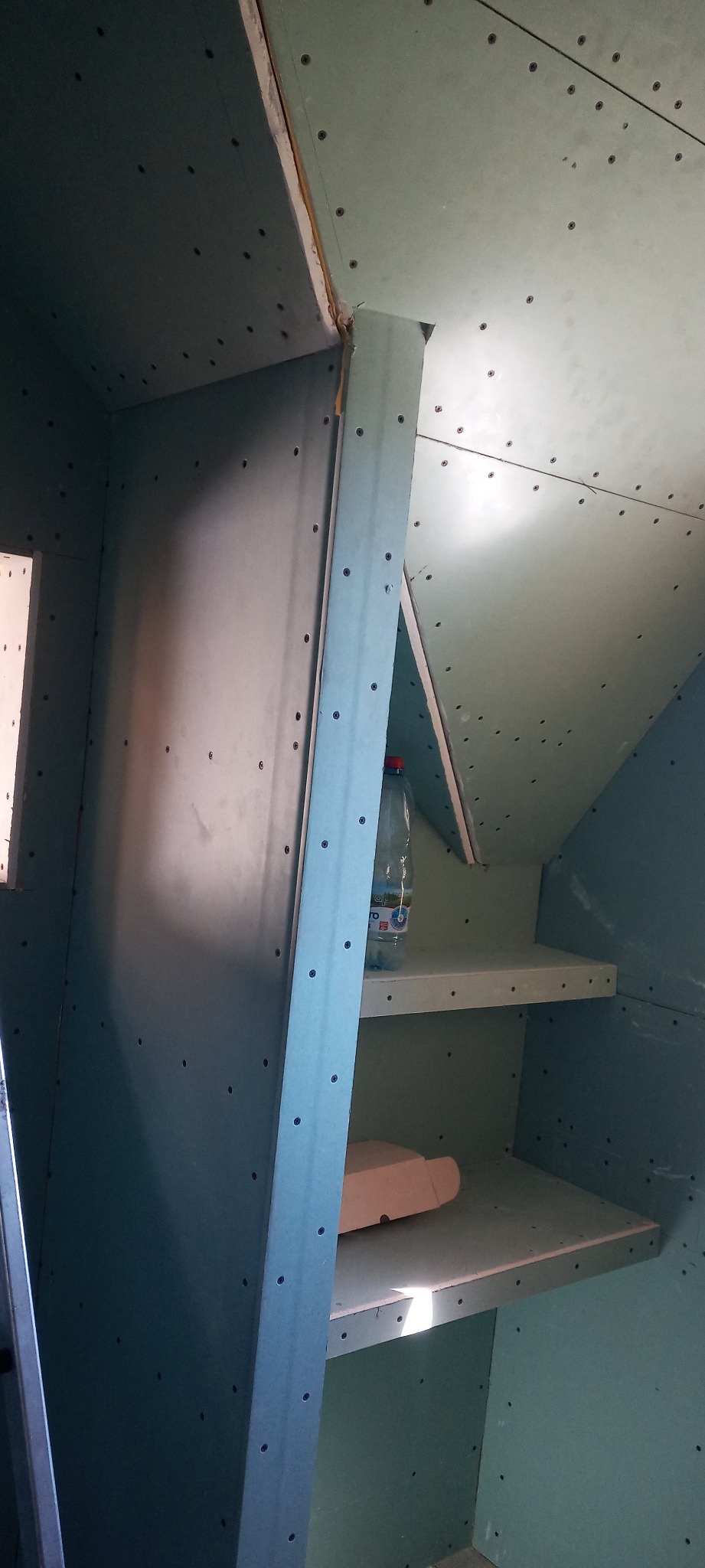 Szkielet szafy wnękowej z płyt kartonowo-gipsowych, widoczne wkręty, metalowy profil i butelka wody na półce.