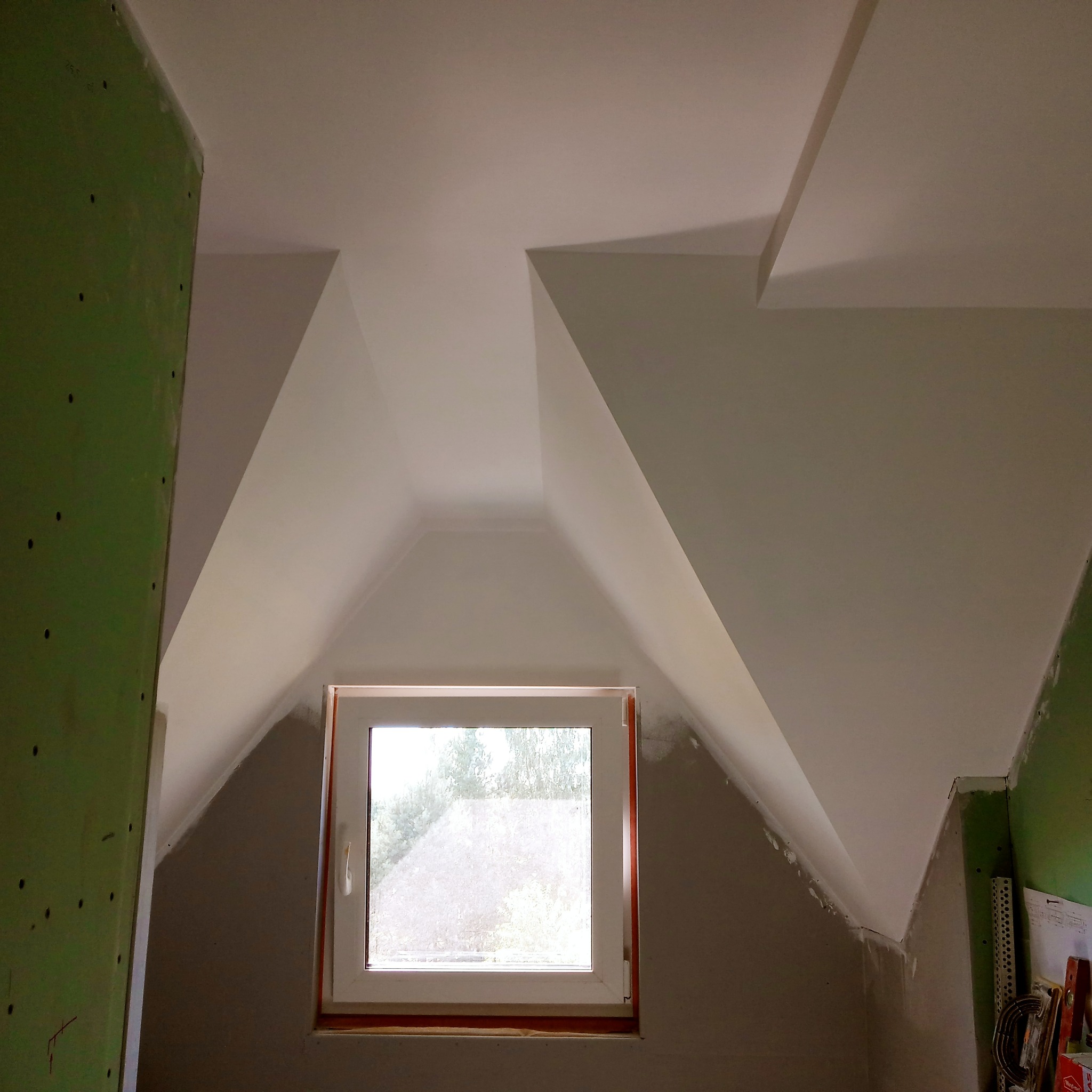 Poddasze z oknem dachowym w trakcie prac wykończeniowych, ściany częściowo pokryte zieloną płytą, sufit i skosy zagruntowane i pomalowane na biało, widoczne niedoskonałości i ślady szpachlowania.